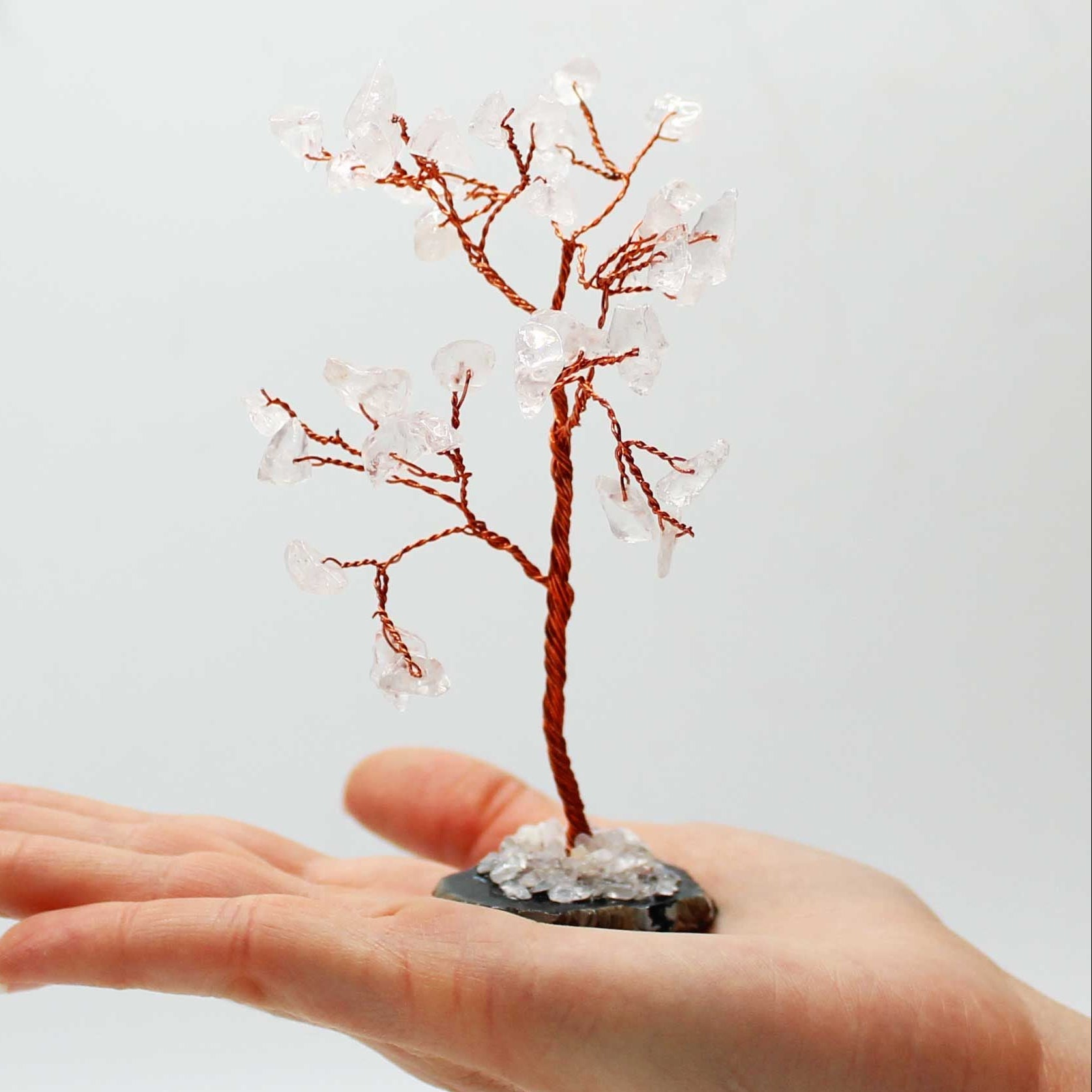 Hand holding a small decorative tree with copper branches and clear beads against a light background