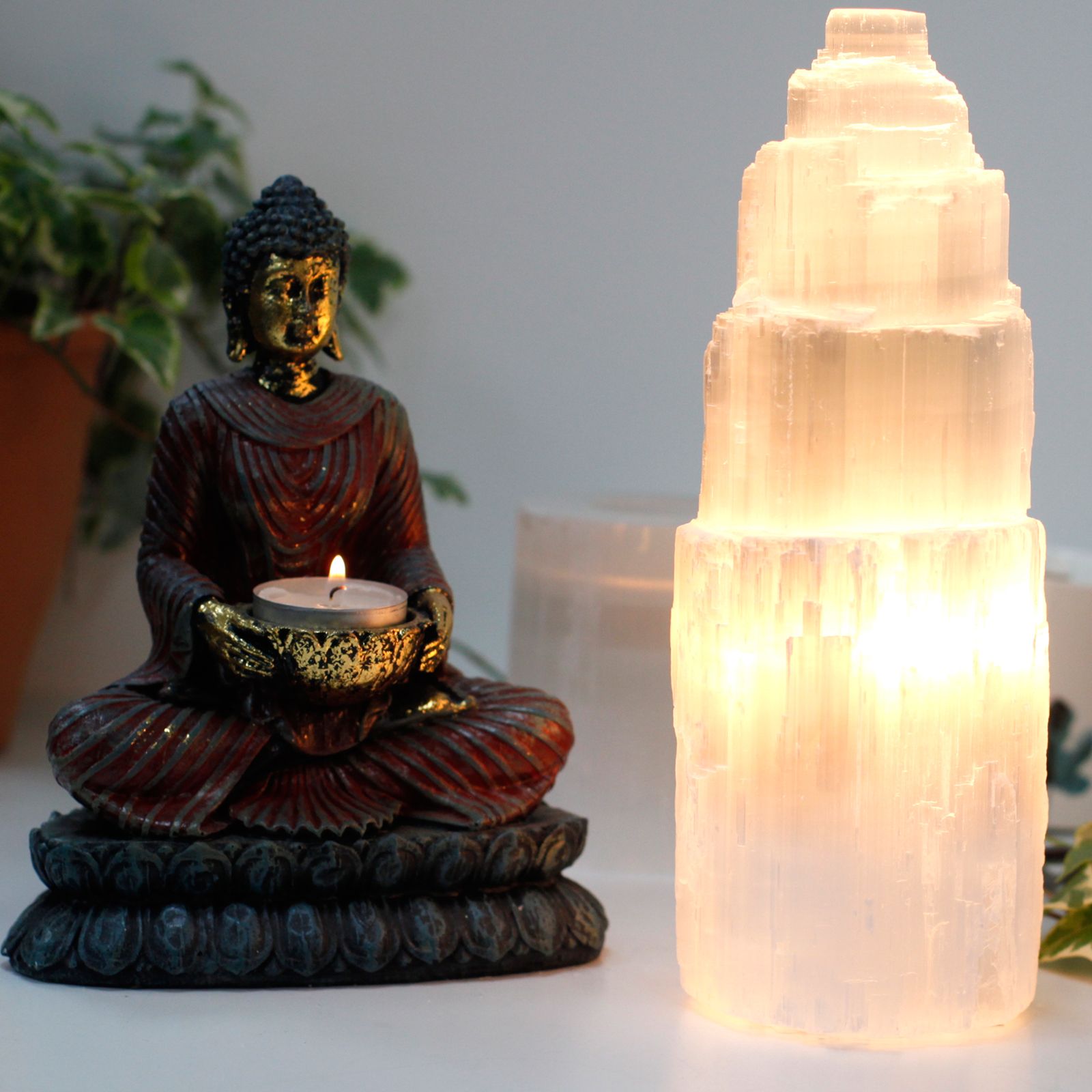 Buddha statue holding a candle next to a selenite tower on a white surface with plants in the background.