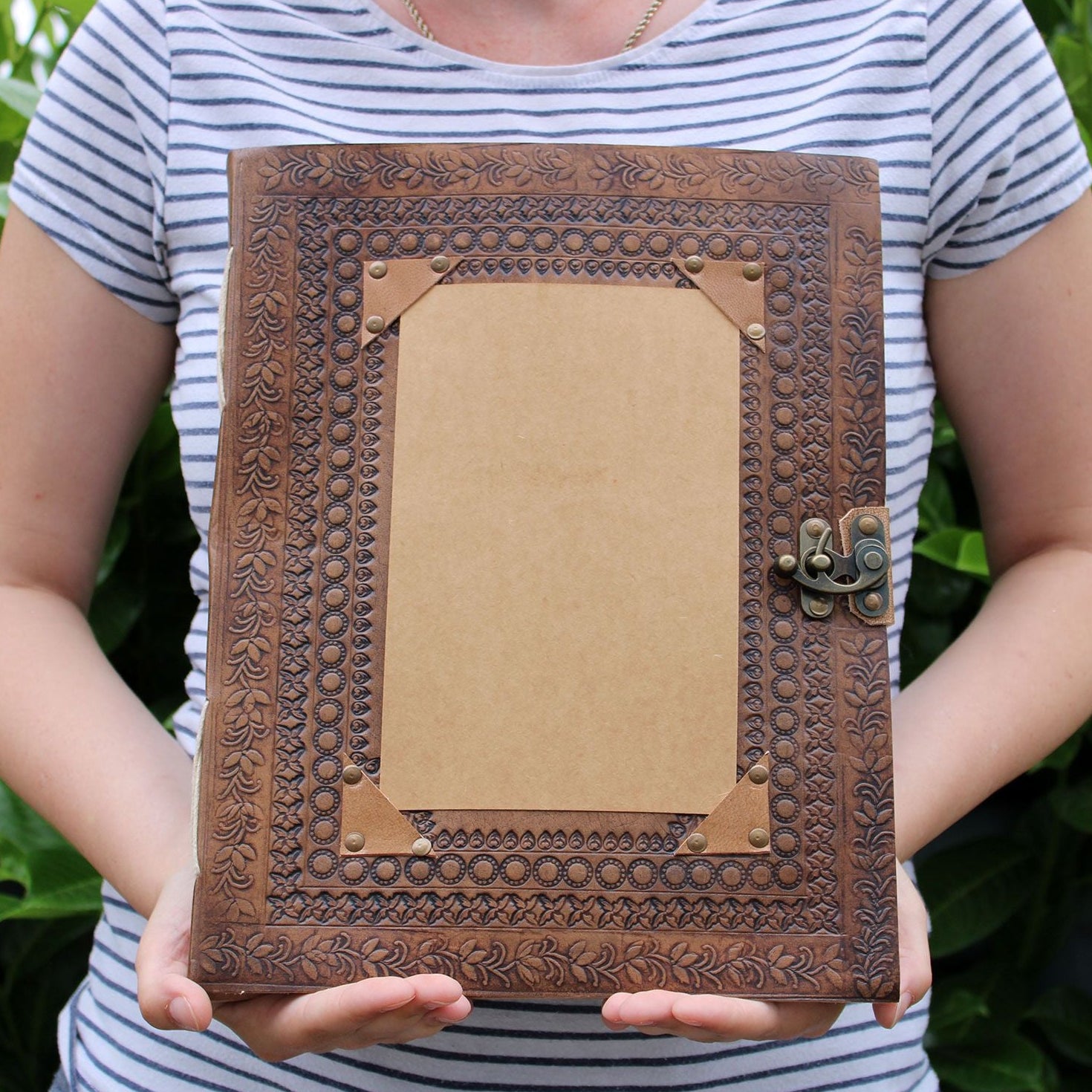 Person holding a decorative brown leather-bound book with intricate patterns against a green leafy background