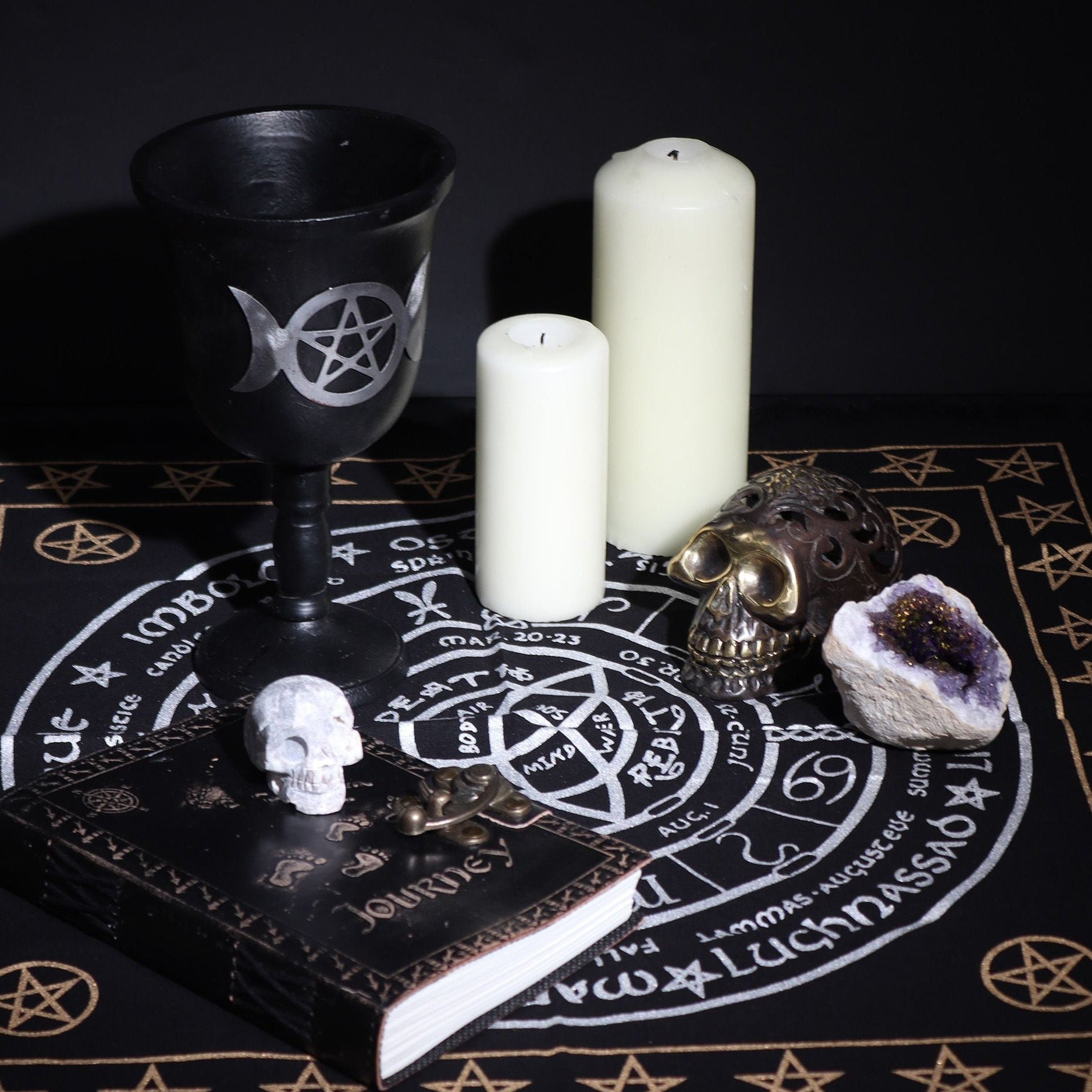 Witchcraft altar setup with black goblet, candles, skull, and crystal on a black cloth with white symbols.