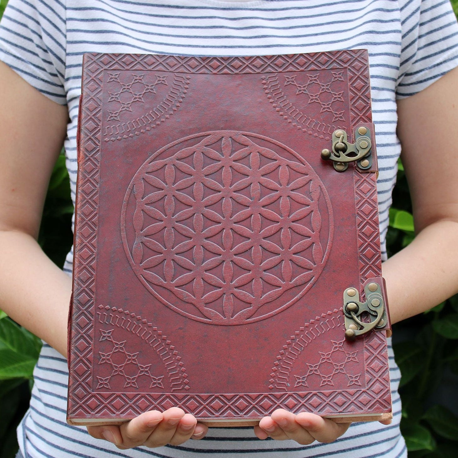 Person holding a red leather notebook with embossed design and metal clasp against a green leafy background