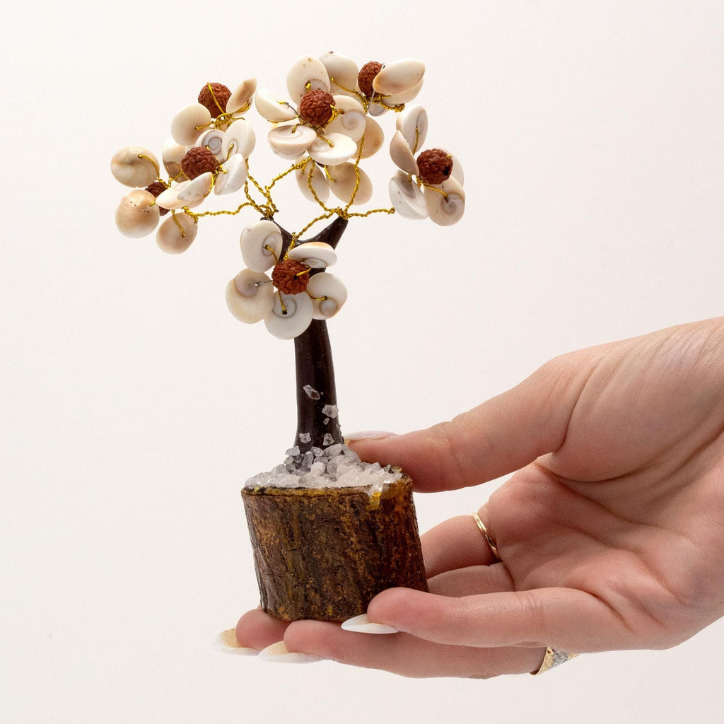 Hand holding a small decorative tree with white flowers and brown trunks against a light background