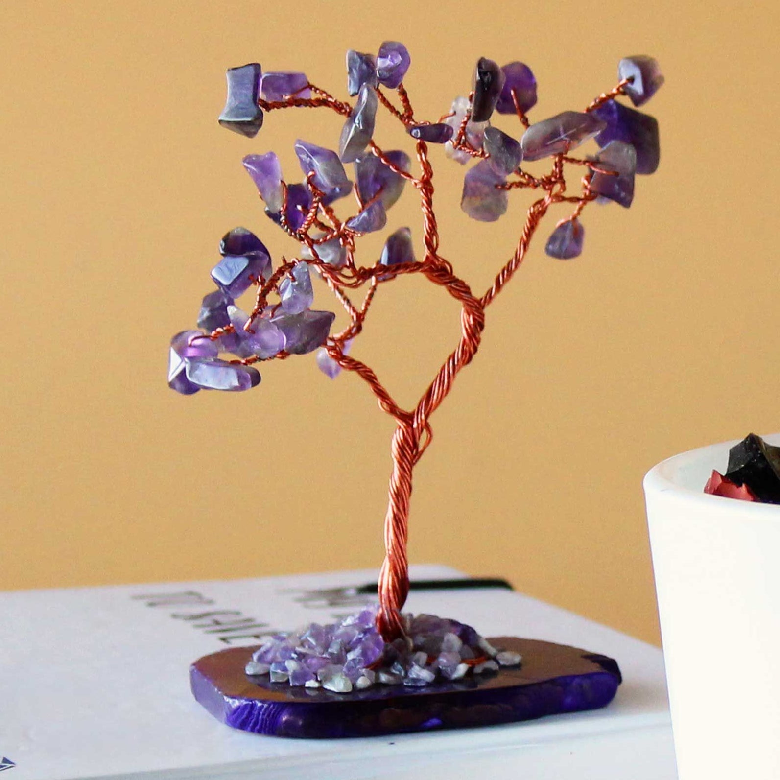 Decorative tree made of copper and purple stones on a book with a white container in the background.